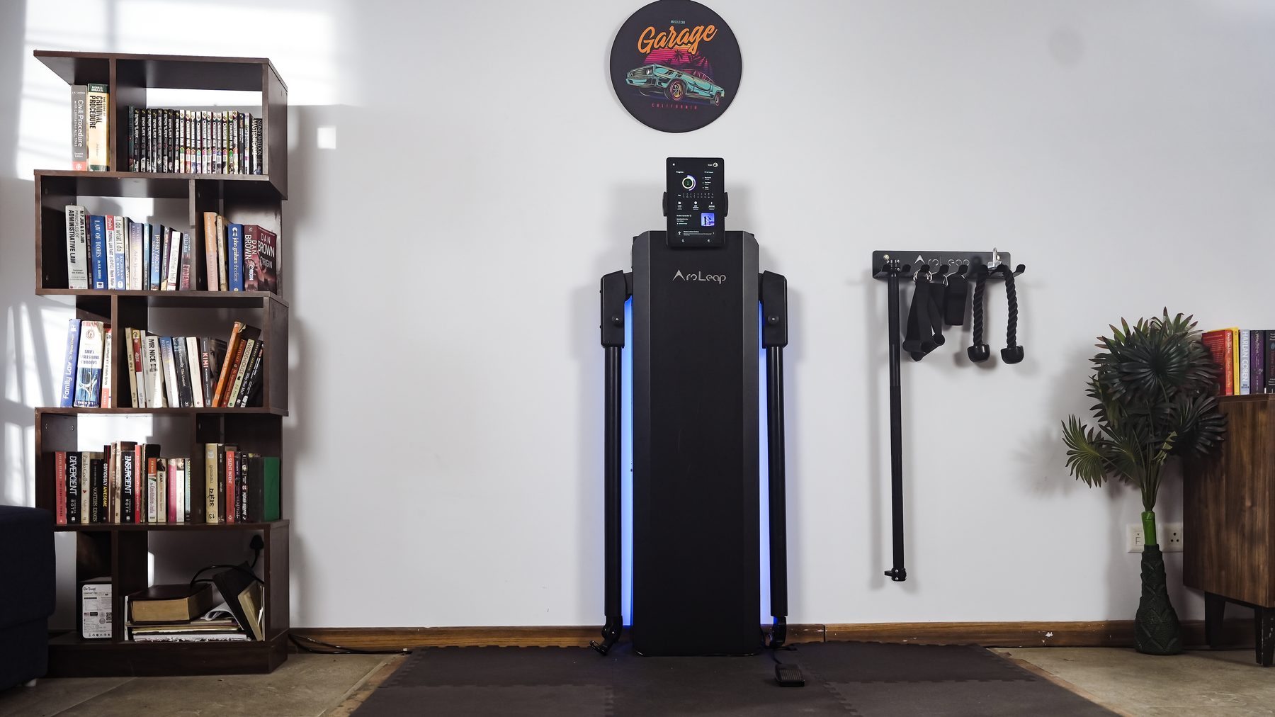 Aroleap X mounted flat against a wall with its accessory hanger, barbell, handles and rope beside it